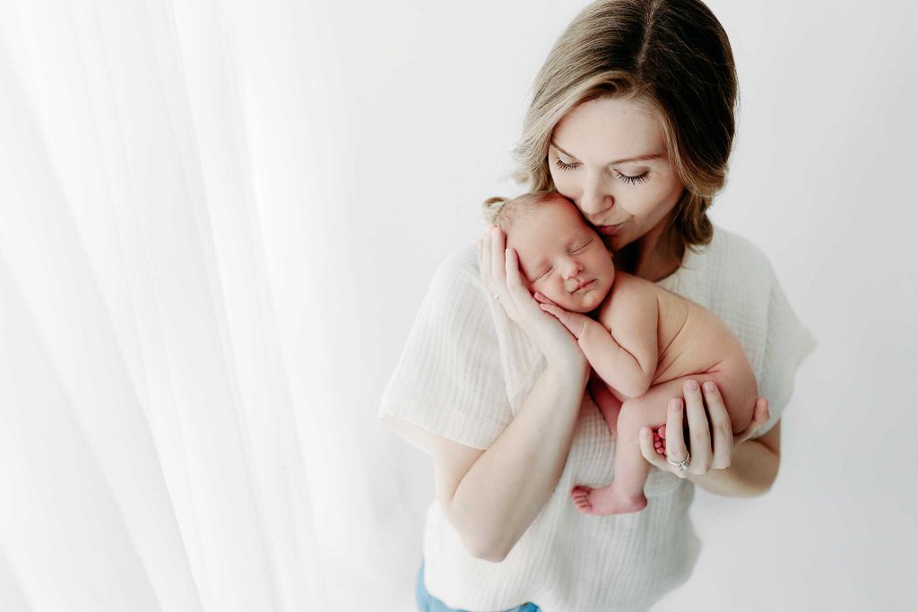 mom kisses naked newborn boy by white window. Baby posed up up in moms arms
