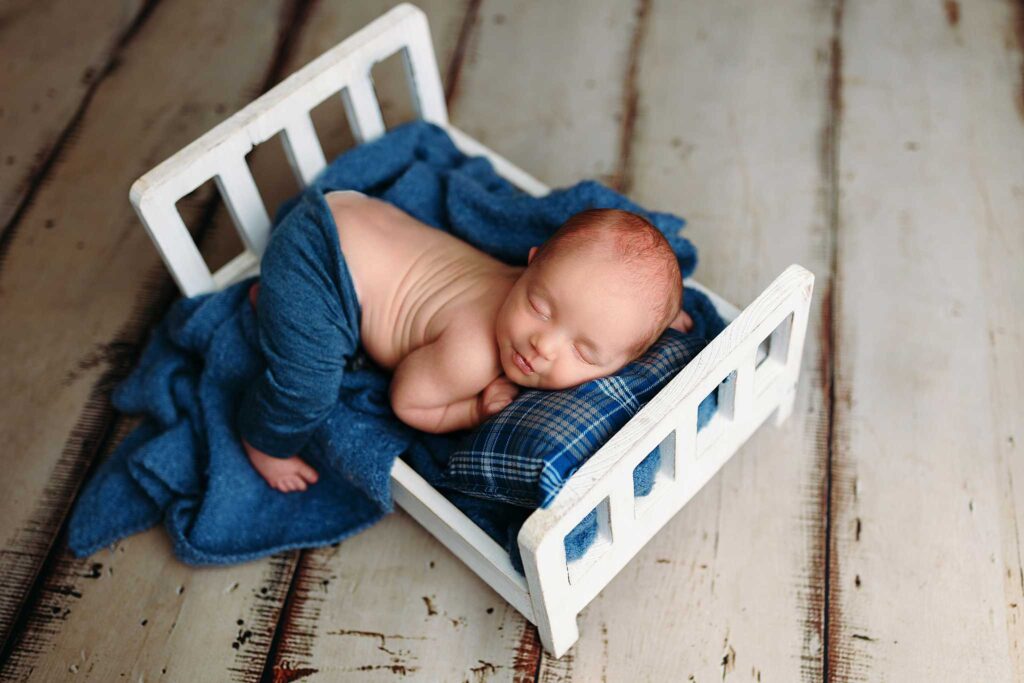 newborn in blue pants hanging off white bed on cream wood