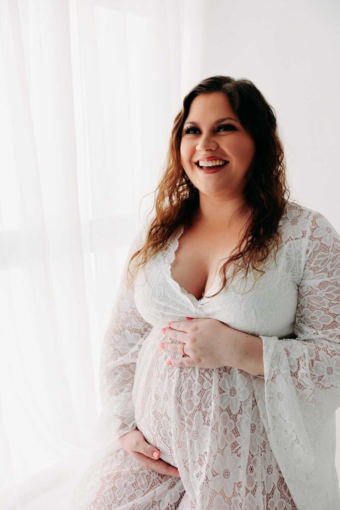 pregnant mom in lace gown smiling by window on white backdrop